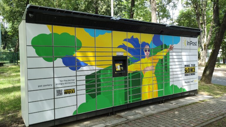 Paczkomat InPost na tle parku, z grafiką kobiety w żółtym stroju na drzwiach skrytek i napisem "Mnie też Słońce dodaje energii".