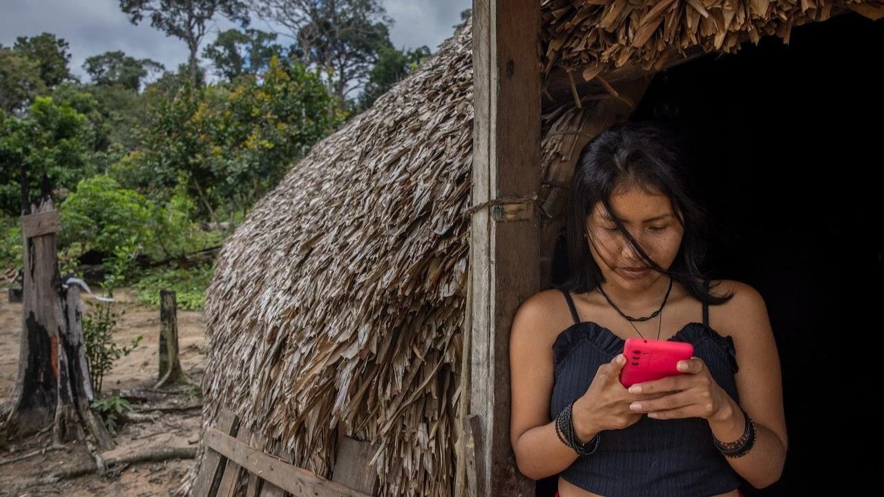 Kobieta z telefonem komórkowym stojąca przed chatką krytą strzechą wśród drzew.