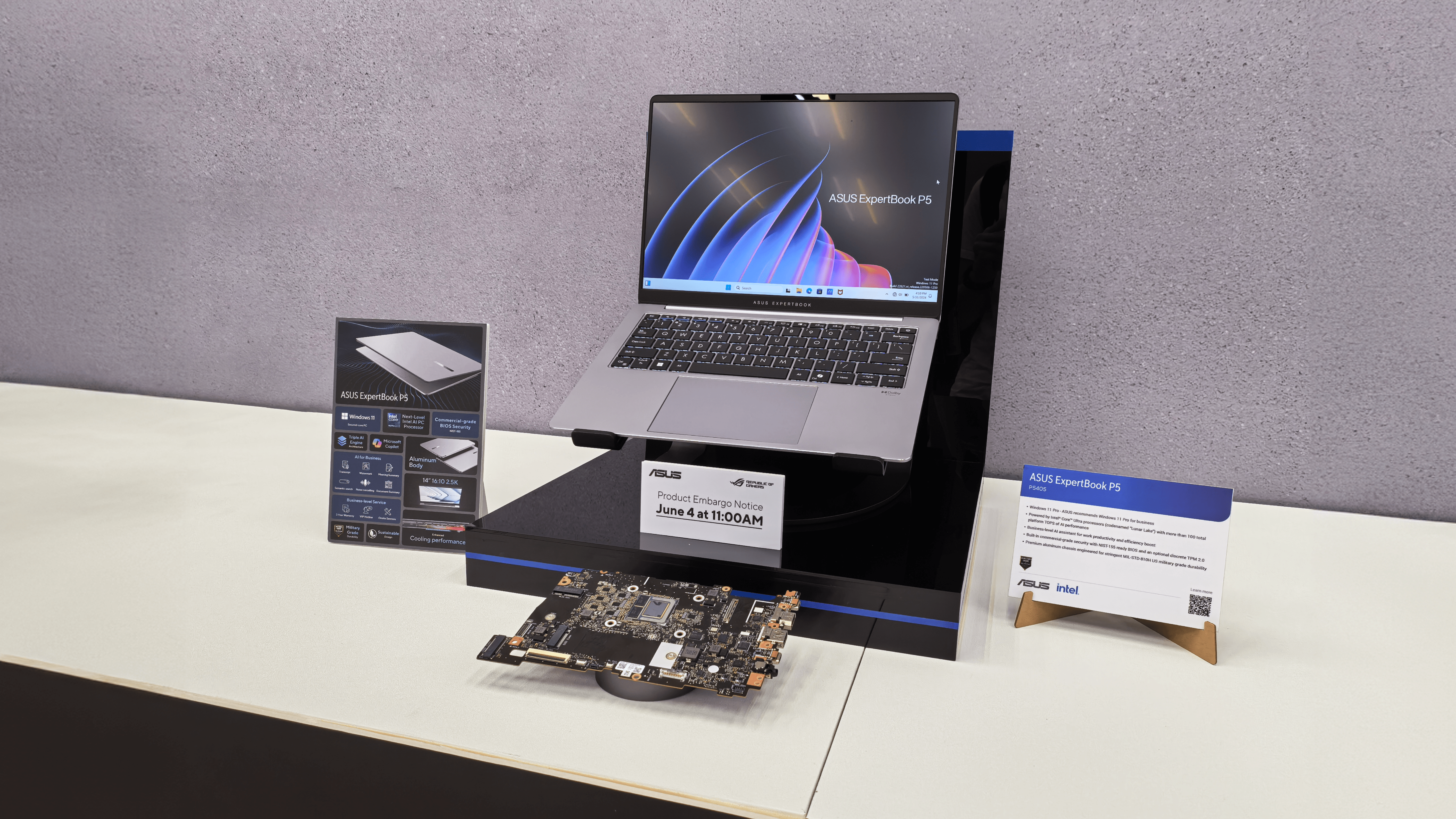 Laptop ASUS ExpertBook P5 na czarnym stojaku na białym stole, z tabliczką informacyjną dotyczącą specyfikacji technicznej i datą embargo produktowego.