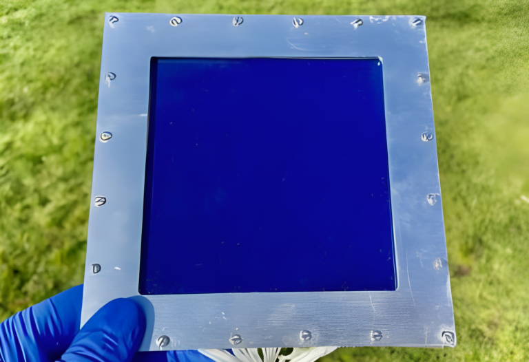 Osoba w niebieskich rękawicach trzyma liście fotowoltaiczne, jeden prototypowy panel z niebieskim szkłem i metalową ramką z śrubami, na tle trawy.