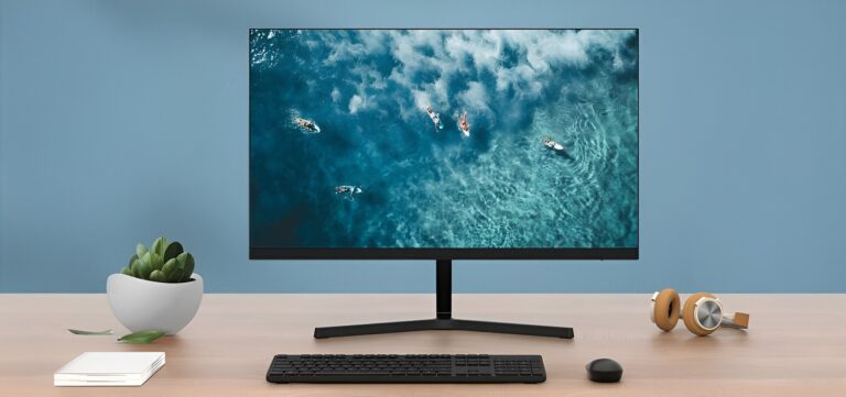 Biurko z monitorem Xiaomi, na którym wyświetlany jest obraz morza z surferami, klawiaturą, myszką, słuchawkami i doniczką z rośliną.