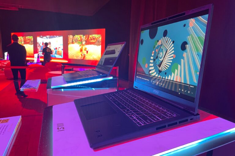 Laptopy MSI na stołach wystawowych podczas eventu w oświetlonym pomieszczeniu z ekranami w tle.