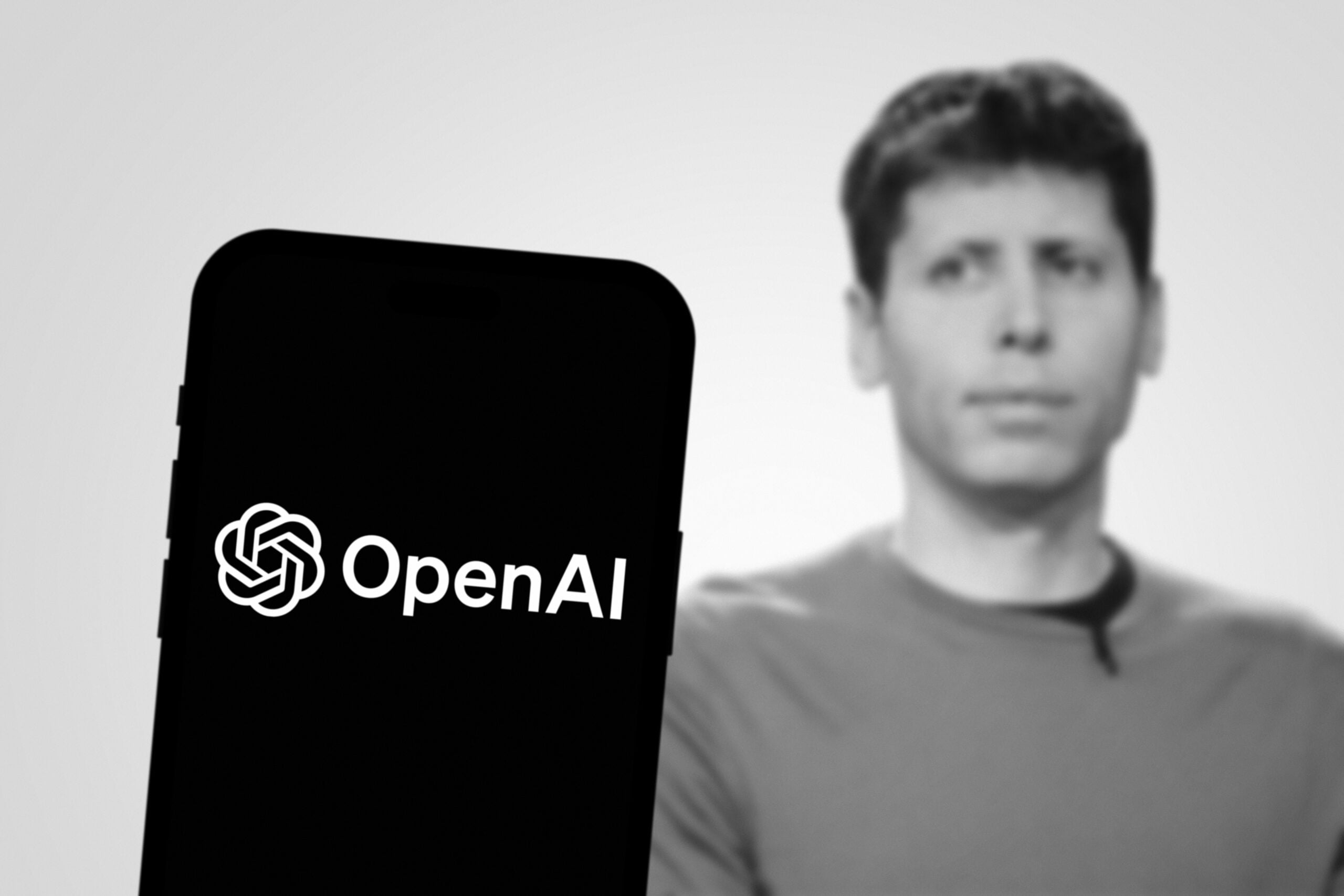 Logo OpenAI na ekranie telefonu komórkowego, w tle niewyraźny wizerunek mężczyzny.