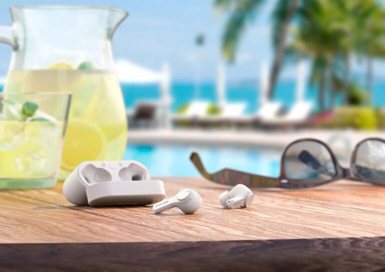 Słuchawki Philips na drewnianym stole, obok dzbanek z lemoniadą i okulary przeciwsłoneczne, w tle basen i palmy.