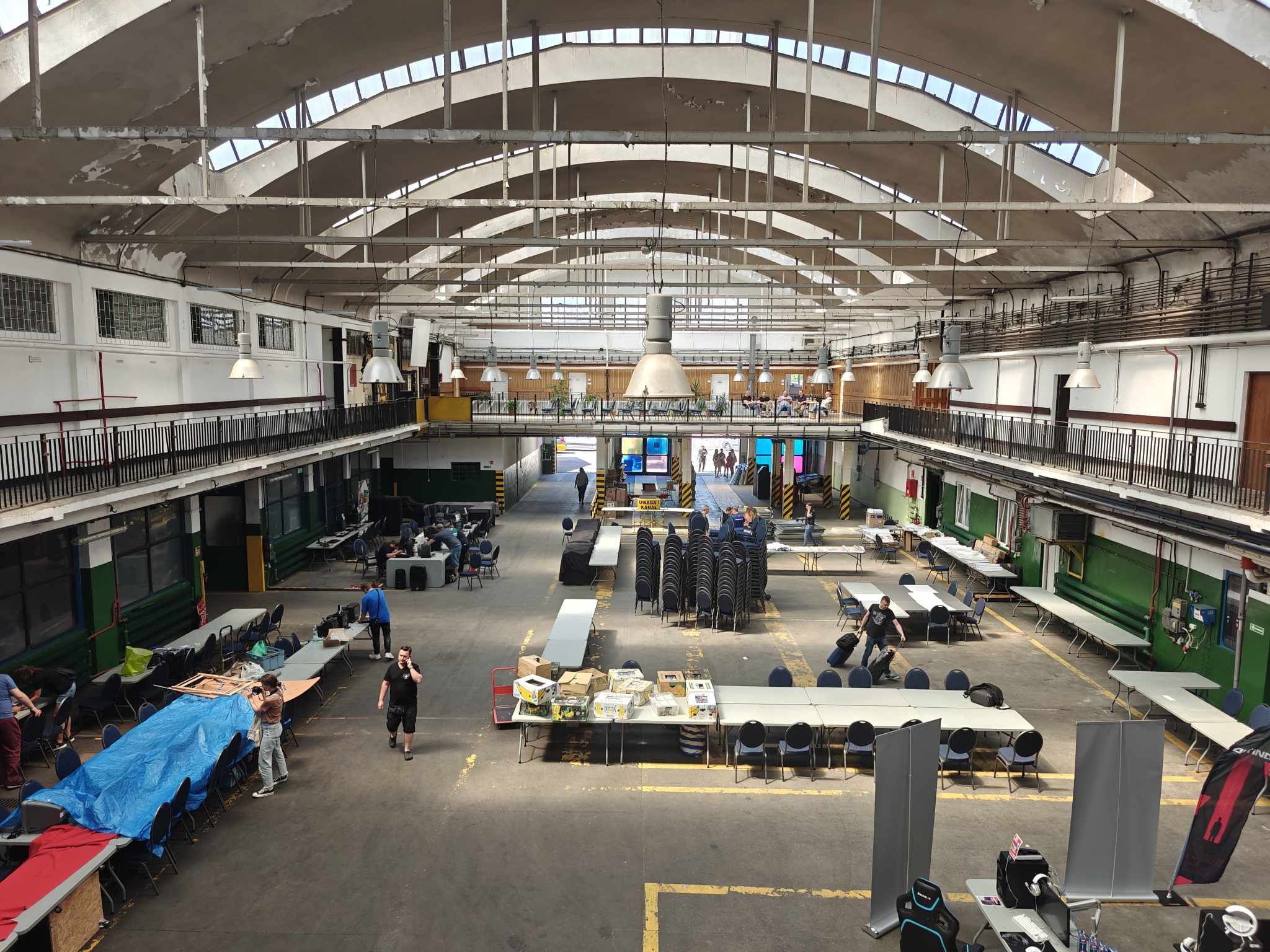 Wnętrze dużej hali przemysłowej z wysokim sklepieniem, stołami, krzesłami i ludźmi przygotowującymi się do wydarzenia Pixel Heaven 2024.