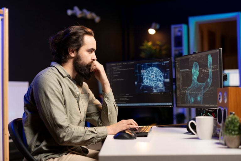 Mężczyzna pracujący przy biurku z dwoma monitorami wyświetlającymi dane naukowe w ciemnym pomieszczeniu. Zarobki w IT są dla niego wysokie