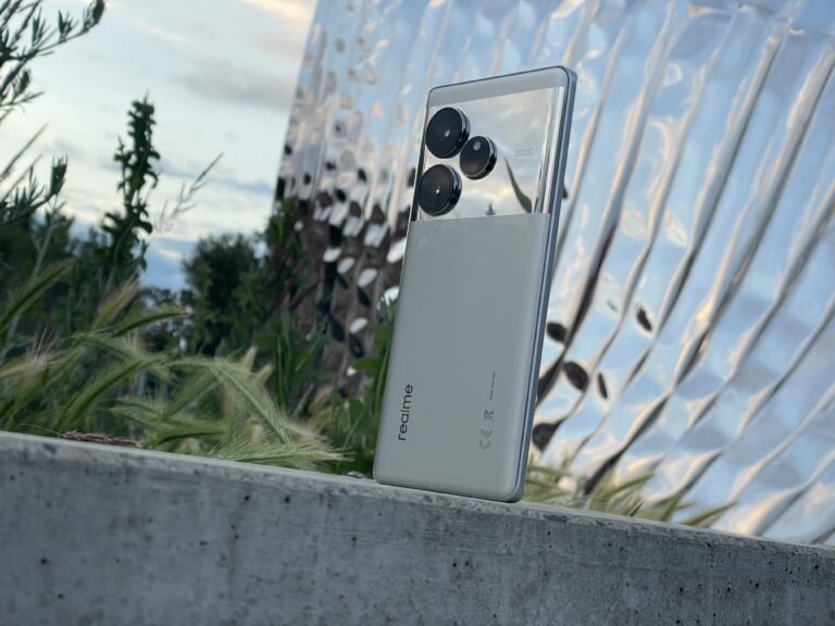Smartfon realme z trzema aparatami fotograficznymi na tle roślin i falowanego metalowego panelu.