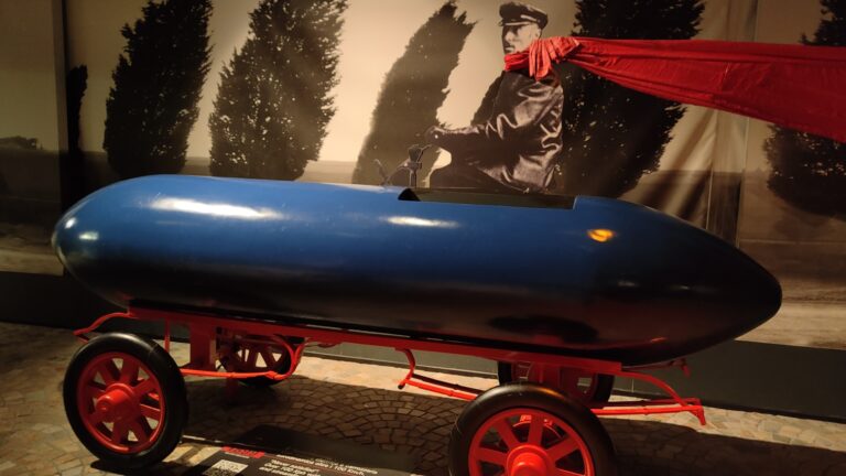Niebieski samochód elektryczny La Jamais Contente na czerwonych kołach w muzeum, w tle czarno-białe zdjęcie mężczyzny z czerwoną szarfą na szyi.
