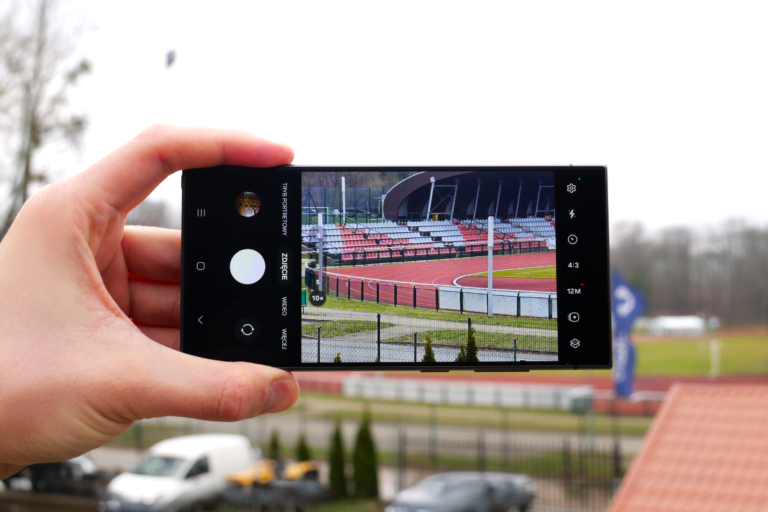 Ręka trzymająca smartfon, na ekranie którego widać zdjęcie stadionu lekkoatletycznego z bieżnią i trybunami.