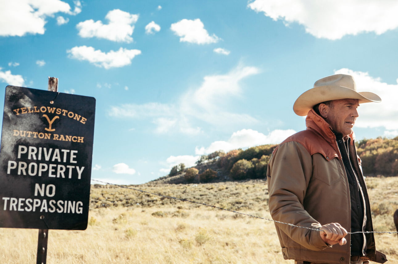 Mężczyzna w kapeluszu kowbojskim i kurtce stoi na tle znak "Yellowstone Dutton Ranch Private Property No Trespassing" na pastwisku.
