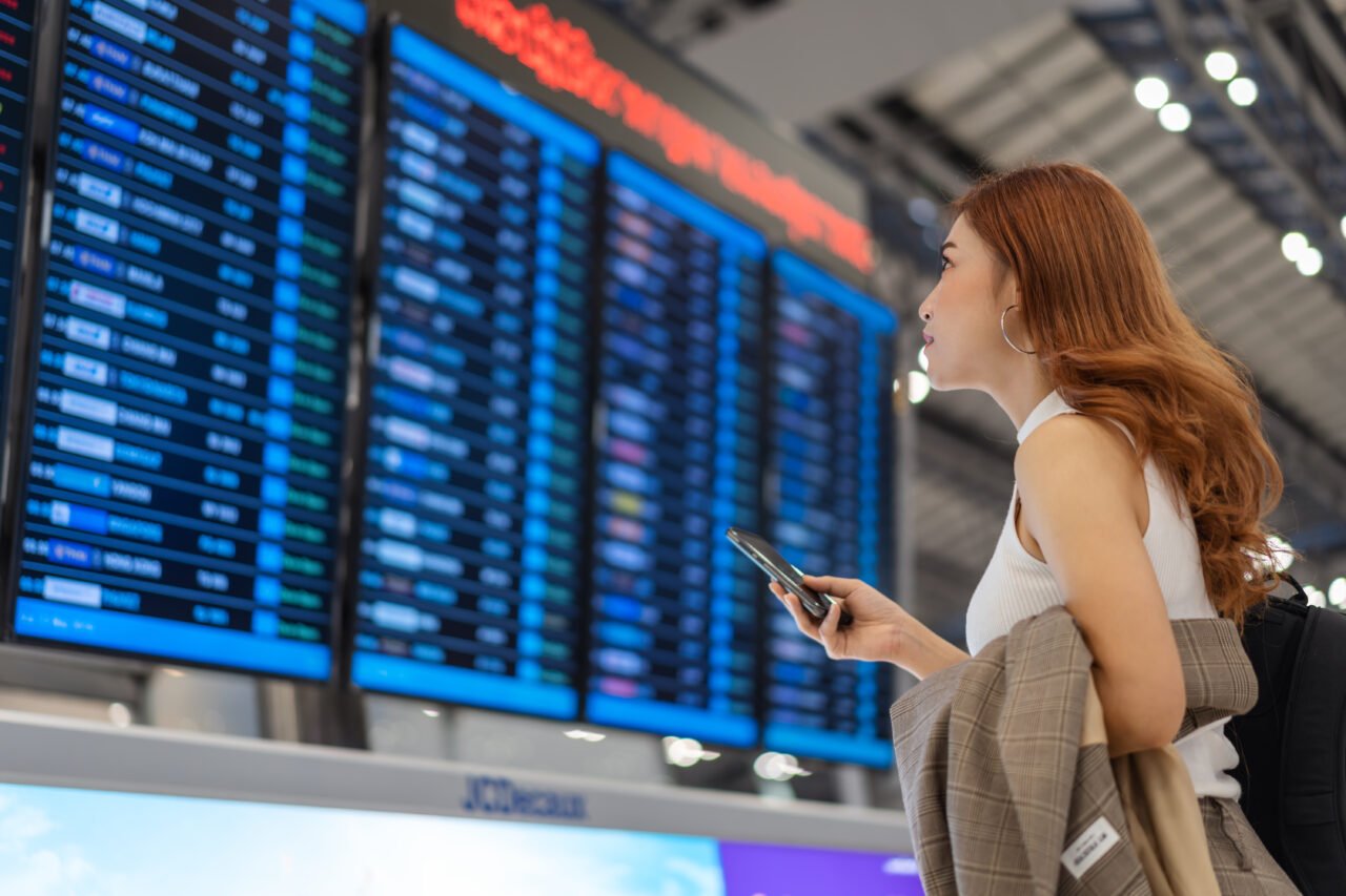 Kobieta patrząca na tablicę odlotów na lotnisku, trzymająca telefon i marynarkę.