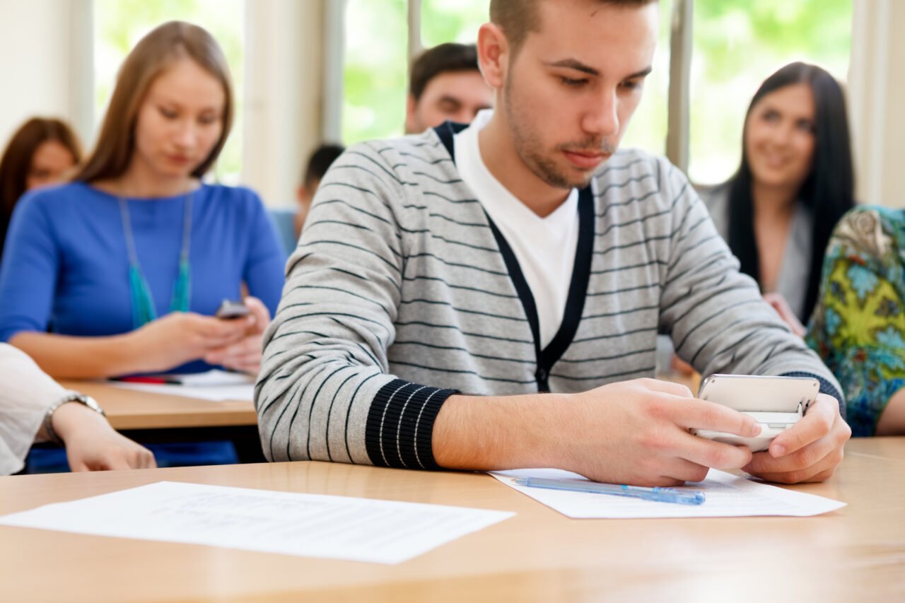 Młody mężczyzna w sali lekcyjnej korzysta z telefonu, w tle grupa studentów.