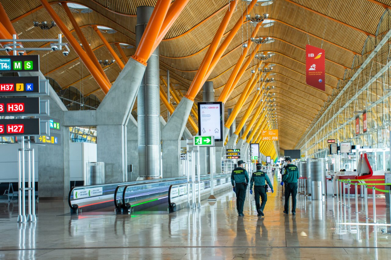 Wnętrze terminalu lotniska z widocznymi oznaczeniami bramek i funkcjonariuszami Guardia Civil patrolującymi halę.