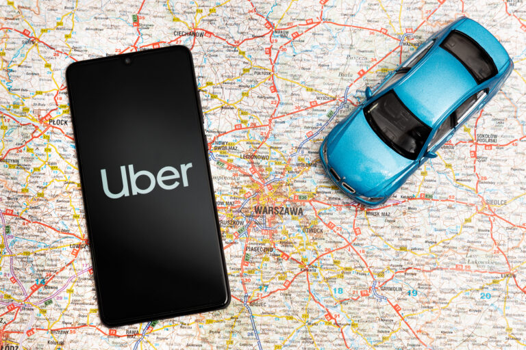 Smartfon z wyświetloną aplikacją Uber oraz niebieski model samochodu na mapie Warszawy.