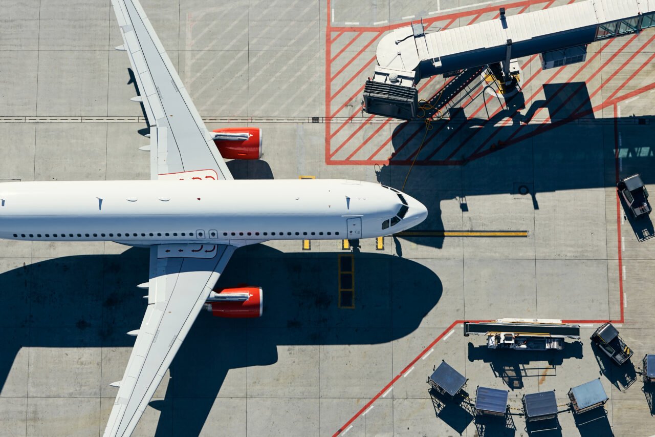 Widok z lotu ptaka na samolot przy gate na płycie lotniska, z rękawem prowadzącym do terminalu i sprzętem lotniskowym wokół samolotu.