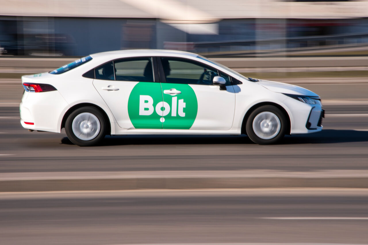 Biała taksówka z logo Bolt na zielonym tle, jadąca po ulicy miejskiej.
