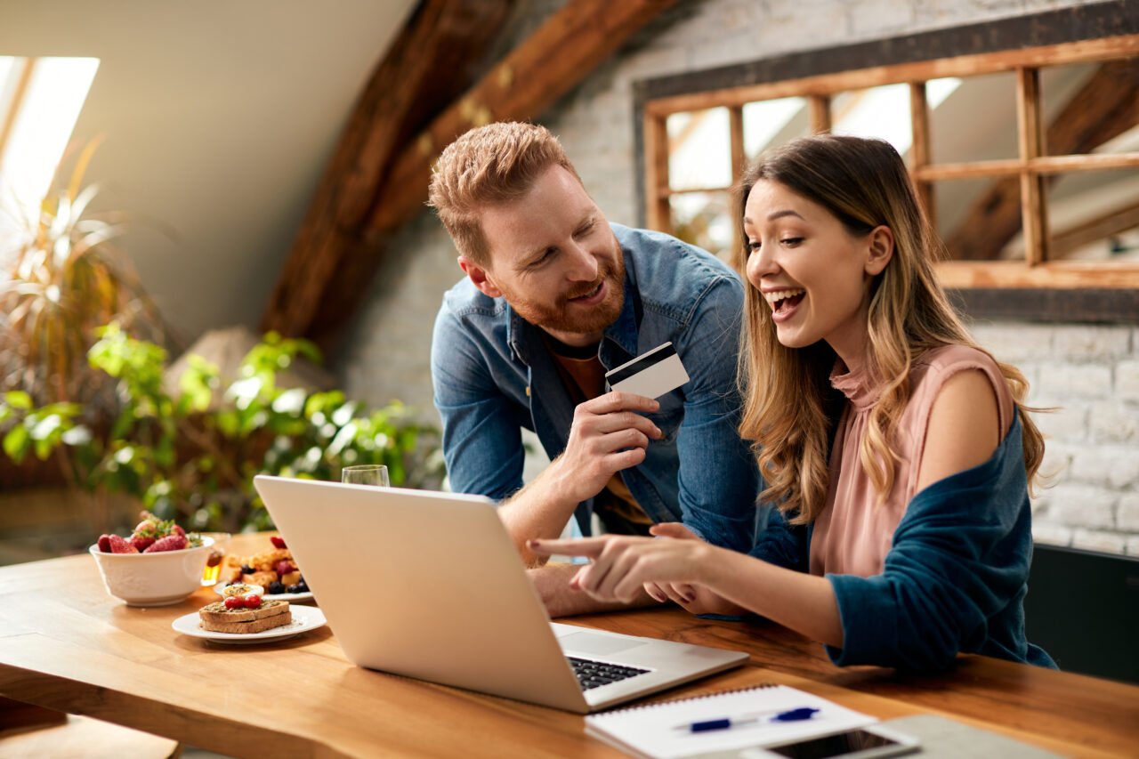 Para korzystająca z laptopa, mężczyzna trzyma kartę kredytową, kobieta wskazuje na ekran i się uśmiecha.