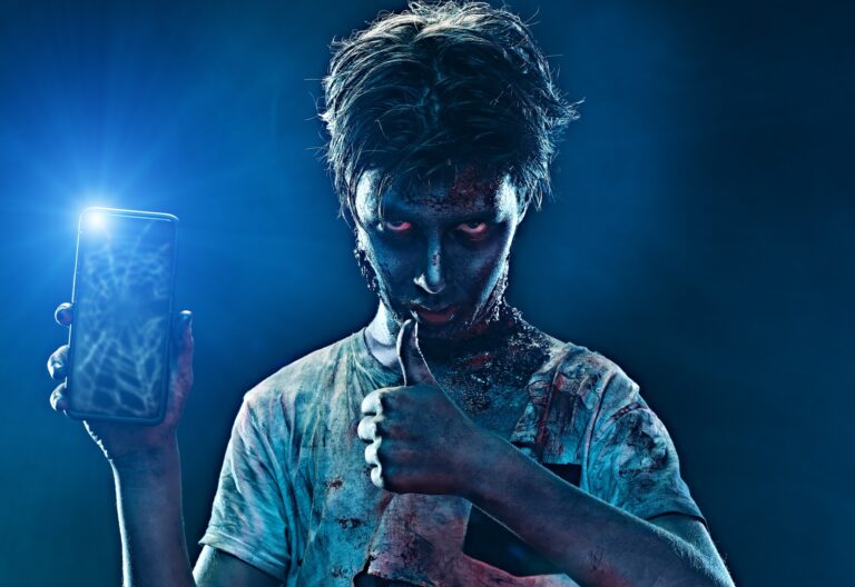 Osoba wyglądająca jak zombie trzymająca smartfon z włączoną latarką i pokazująca kciuk w górę.