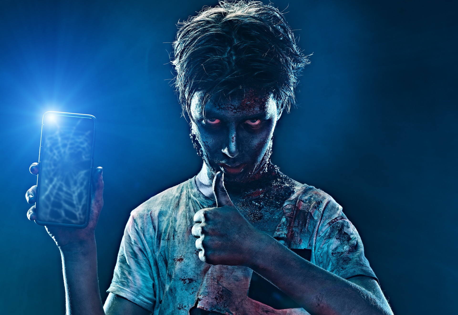 Osoba wyglądająca jak zombie trzymająca smartfon z włączoną latarką i pokazująca kciuk w górę.