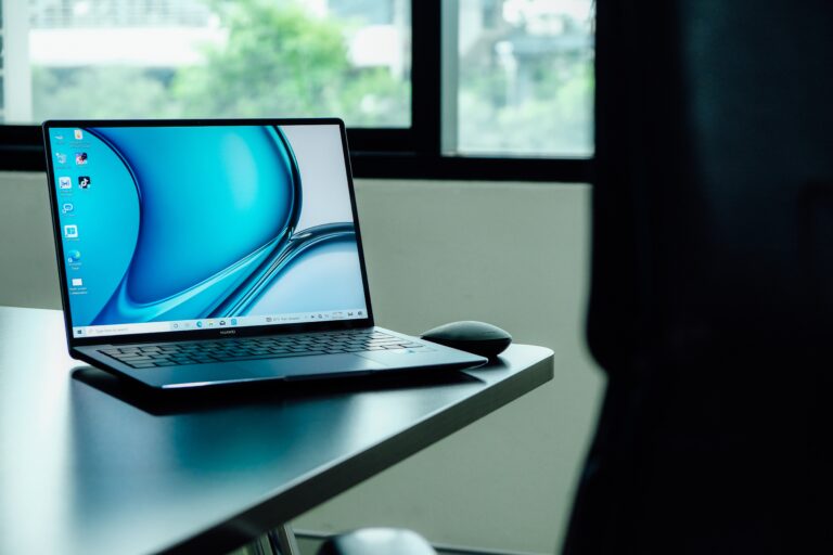 Laptop z otwartym ekranem stojący na biurku, z myszką obok, na tle okna.