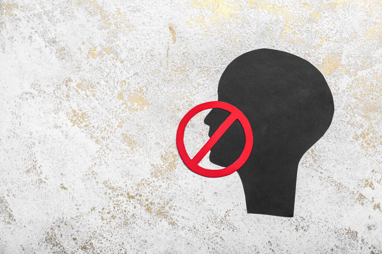 Czarny zarys głowy z czerwonym symbolem zakazu wokół ust na tle szarego marmuru z złotymi plamkami.