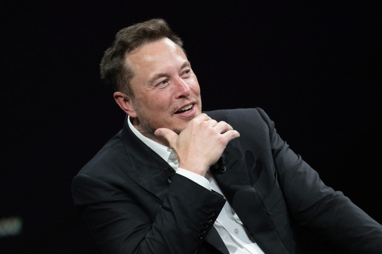Elon Musk. Mężczyzna w garniturze, trzymający rękę przy brodzie, siedzący na ciemnym tle.