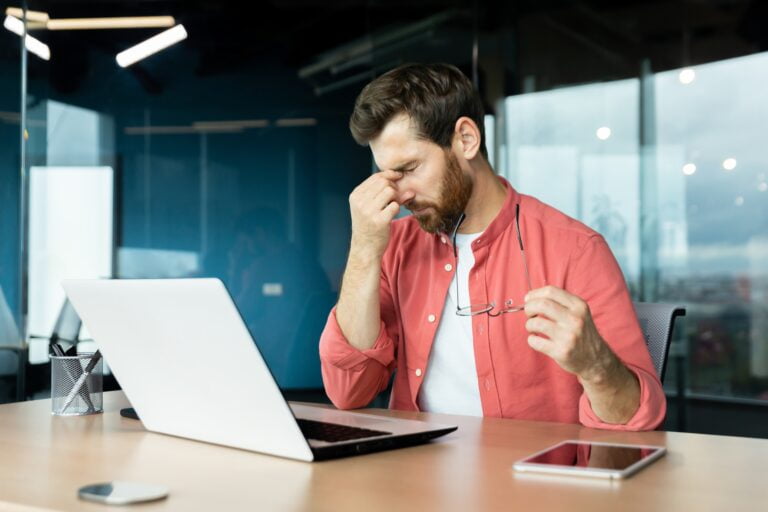 Mężczyzna trzymający okulary, pocierający nos, siedzący przy biurku z laptopem.