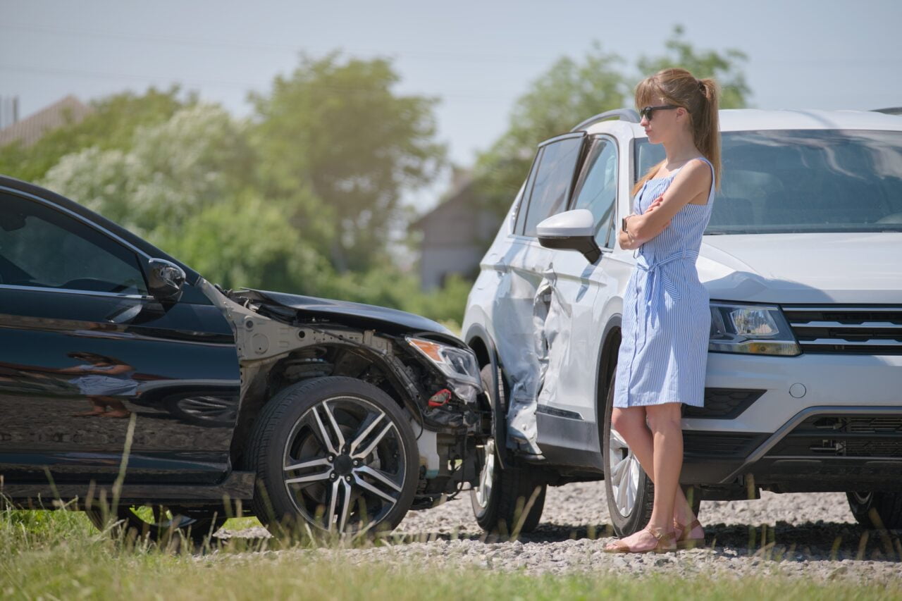 Kobieta w okularach przeciwsłonecznych stojąca przy dwóch uszkodzonych samochodach po kolizji.