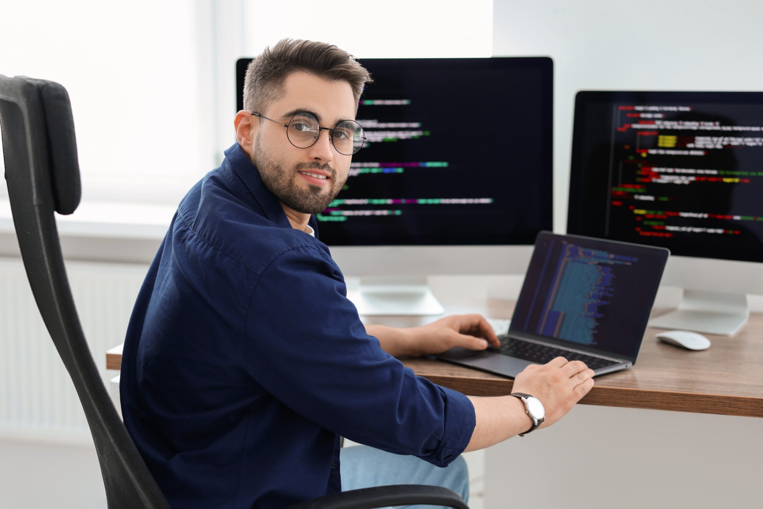 Mężczyzna siedzący przy biurku, pracujący na laptopie z ekranem pełnym kodu, z dwoma monitorami w tle również wyświetlającymi kod.