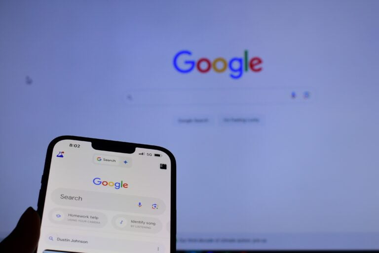 Ekran smartfona z otwartą aplikacją Google oraz w tle strona główna wyszukiwarki Google na ekranie laptopa.
