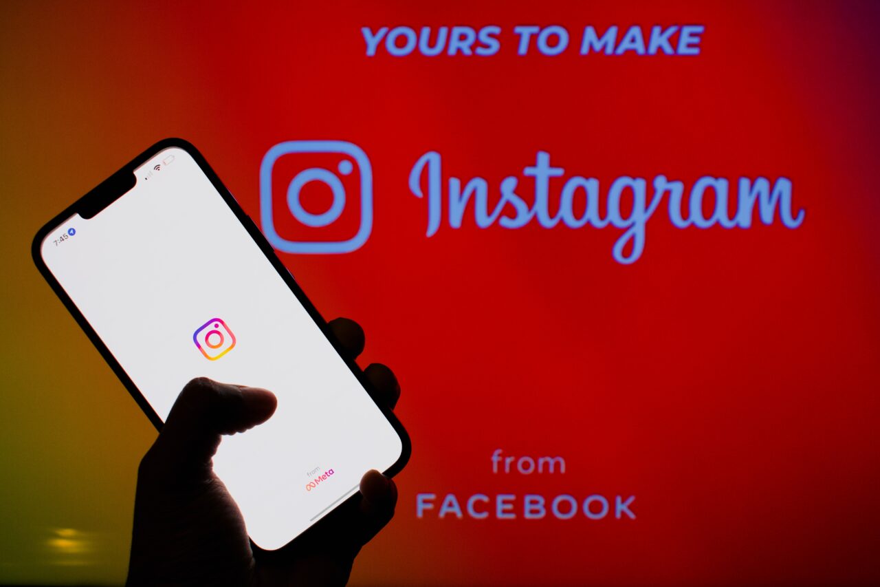 Osoba trzymająca smartfon z logo Instagram na ekranie, w tle widoczne jest powiększone logo Instagram na czerwonym tle oraz napis "YOURS TO MAKE" i "from FACEBOOK".