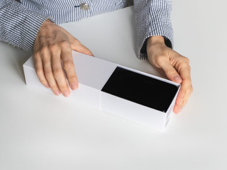 Osoba otwierająca białe pudełko z czarnym smartfonem w środku.
