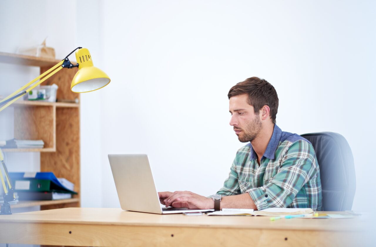 Mężczyzna pracujący przy biurku na laptopie, w tle żółta lampa i półki.