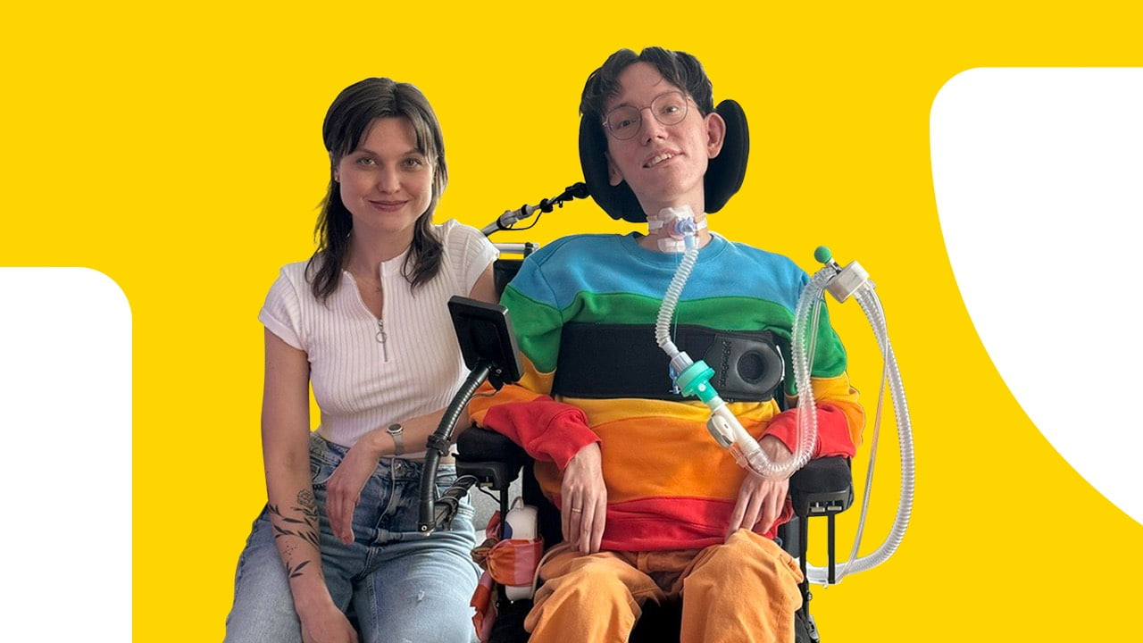 Kobieta w białej koszulce siedzi obok mężczyzny na wózku inwalidzkim z respiratorem, w kolorowej bluzie; tło żółte.