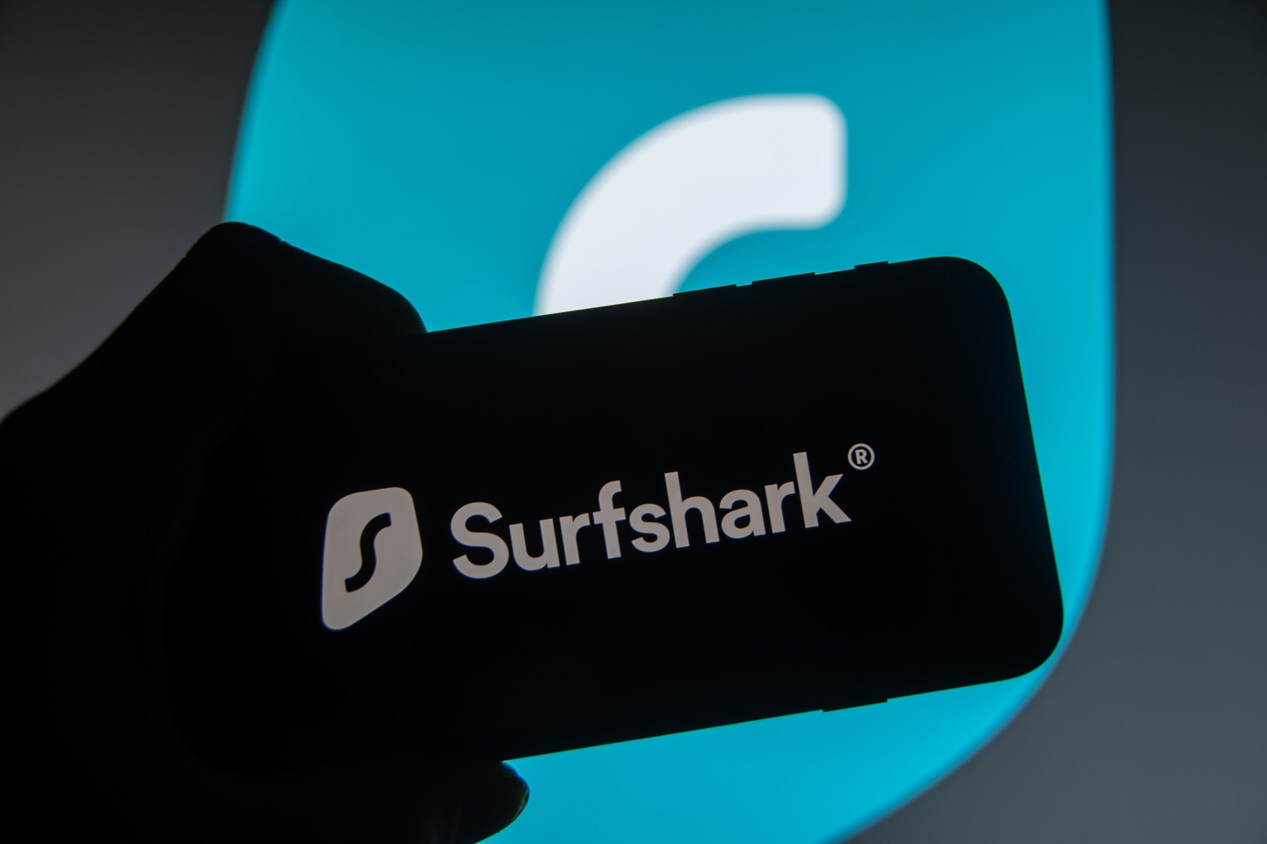 Logo Surfshark VPN cena wyświetlane na ekranie smartfona na tle większej wersji logo.