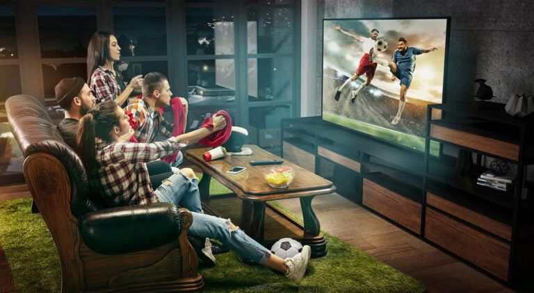 Grupa młodych osób oglądająca mecz piłki nożnej w telewizji, siedząca na kanapie w salonie.
