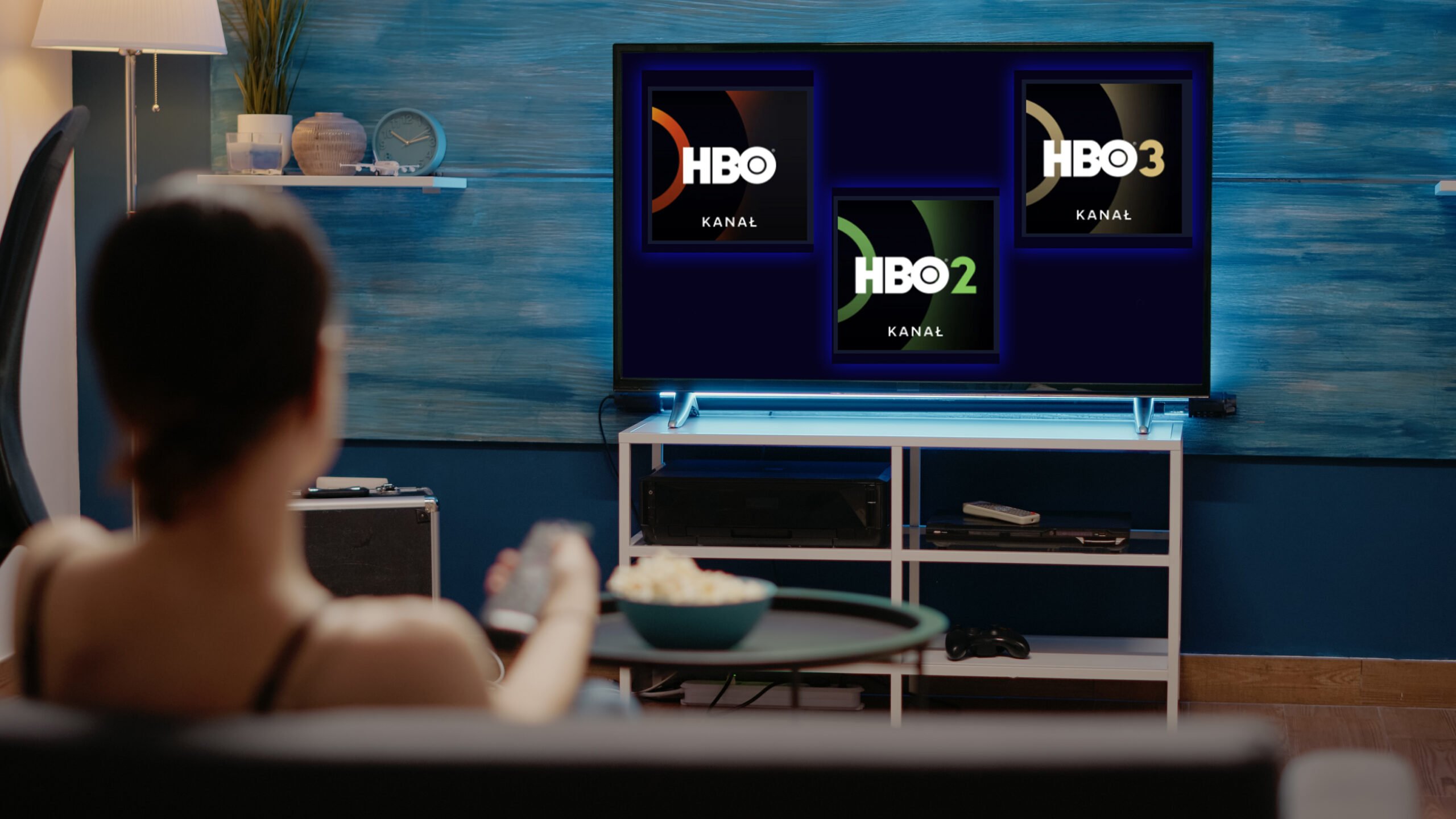 Osoba siedząca przed telewizorem z wyświetlonymi logotypami kanałów HBO, HBO2 i HBO3.