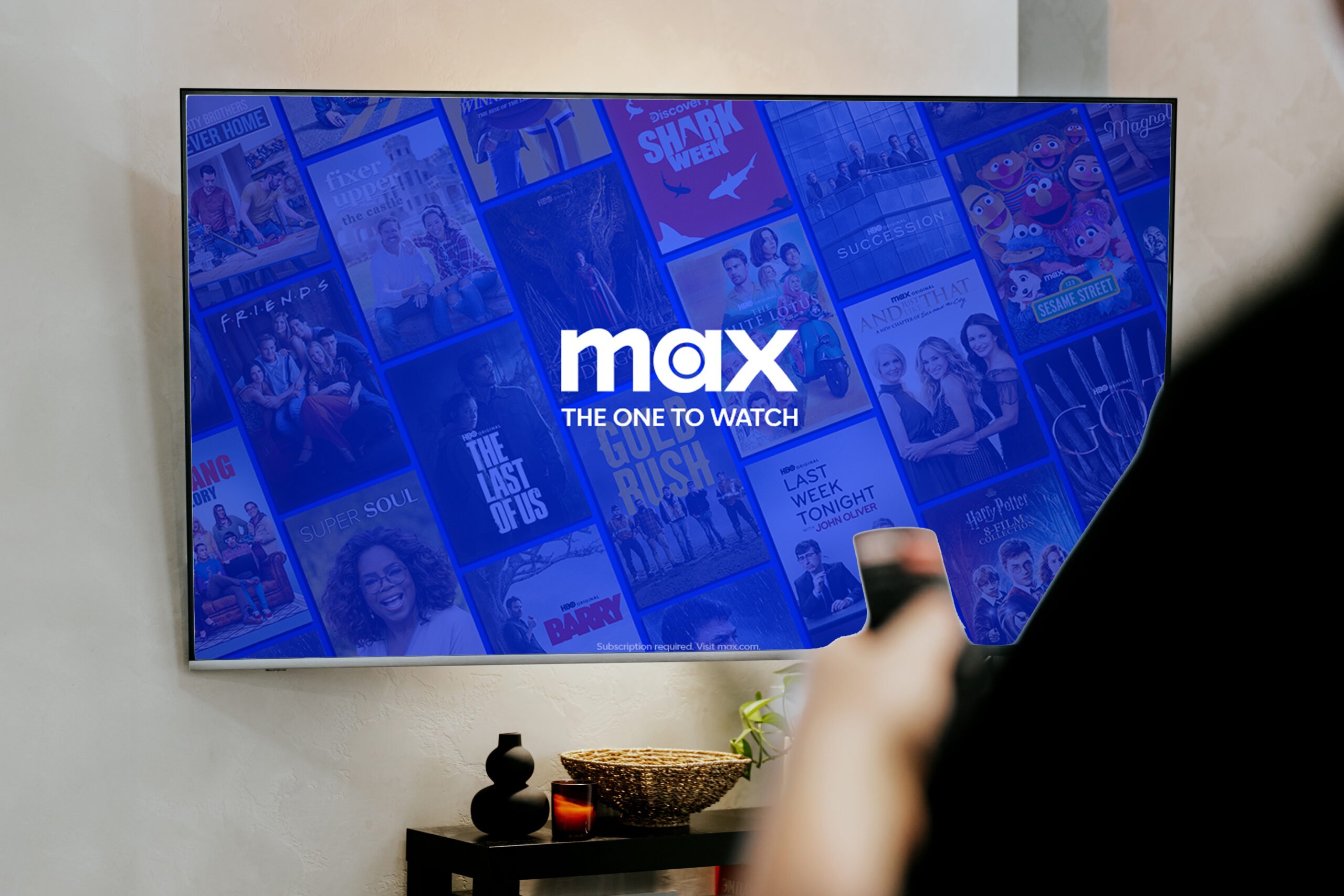 Telewizor z platformą Max w trybie ekranu głównego, wyświetlający różne programy telewizyjne i filmy; w tle widać rękę trzymającą pilot.