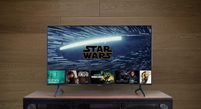 Telewizor wyświetlający logo Star Wars oraz miniaturki powiązanych programów, stojący na drewnianym stoliku przy ścianie pokrytej panelami.