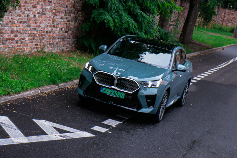 BMW iX2 na drodze w czasie testu, zielony samochód na tle ceglanego muru i zieleni.