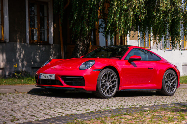 Test Porsche 911 Carrera, czerwony samochód zaparkowany na brukowanej uliczce w cieniu drzew.