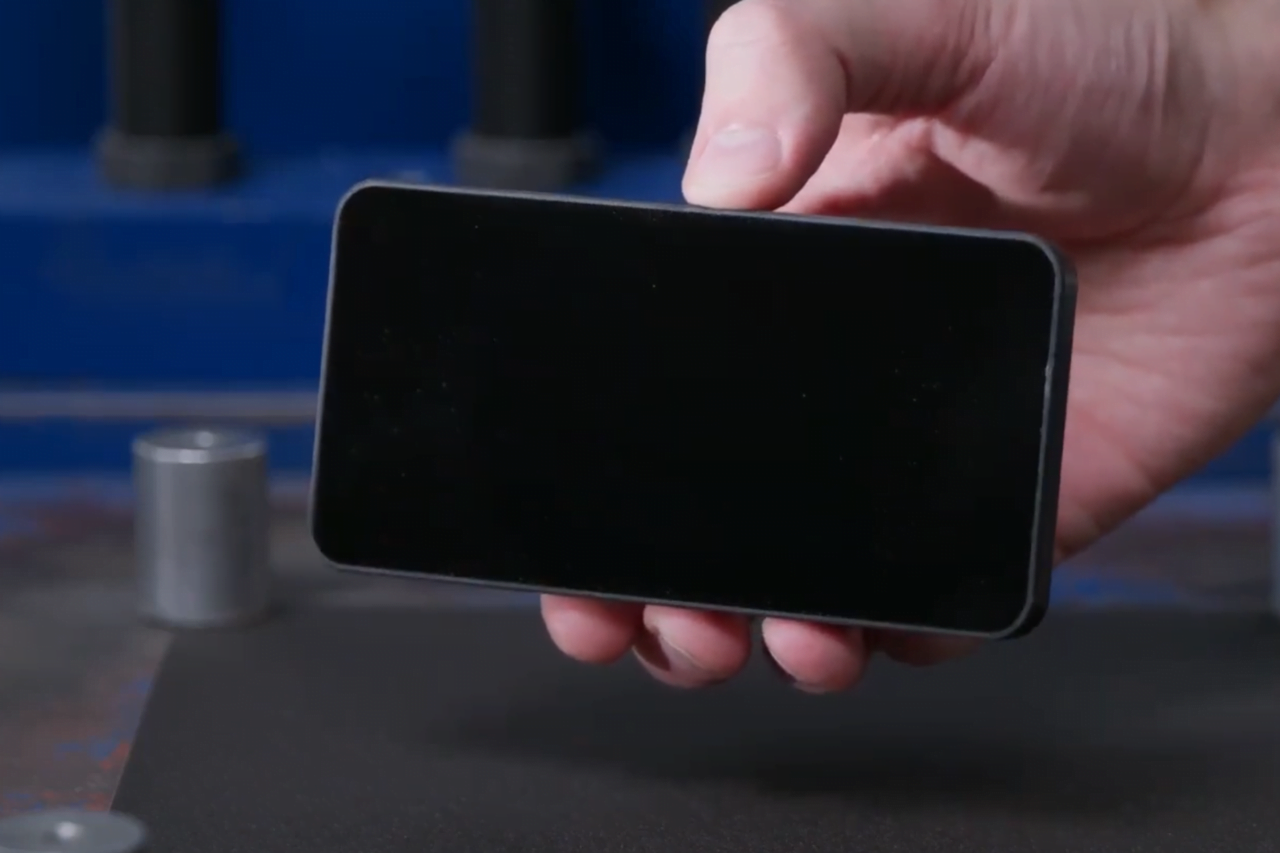 Ręka trzymająca czarny prostokątny przedmiot, najprawdopodobniej smartfon, na tle rozmytej przestrzeni warsztatowej.