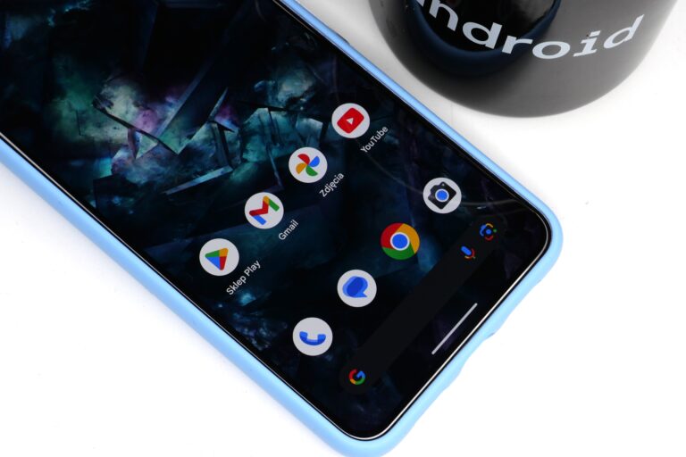 Smartfon z otwartym ekranem głównym, na którym widać ikony aplikacji: Sklep Play, Gmail, Zdjęcia, YouTube, Przeglądarka, Zdjęcia, Telefon oraz Wyszukiwarka Google; obok leży kubek z napisem "Android".