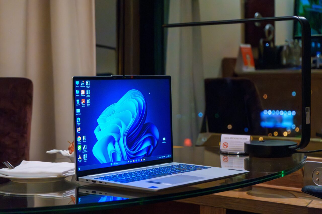 Laptop na biurku z włączonym ekranem systemu Windows 11 oraz nakrycie do posiłku obok.