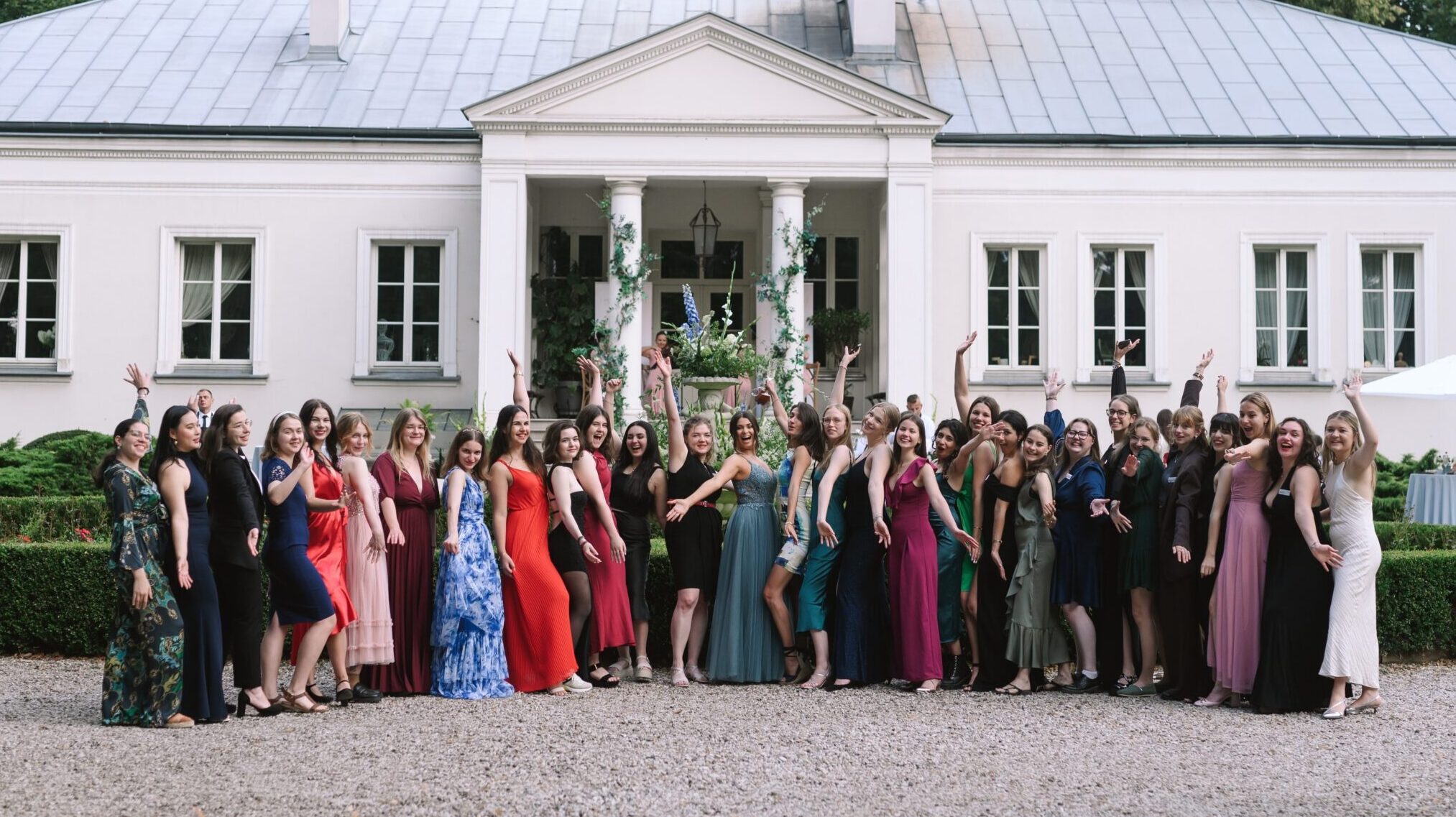 Huawei Summer School for Female Leadership Farewell Gala. Grupa kobiet w eleganckich sukienkach stojąca przed dużym, białym budynkiem z kolumnami i metalowym dachem.