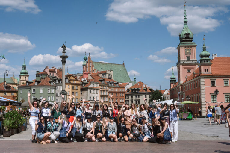 Huawei Summer School for Female Leadership in the Digital Age 2024. Grupa ludzi pozuje do zdjęcia na placu Zamkowym w Warszawie, w tle widoczne są zabytkowe kamienice oraz Zamek Królewski.