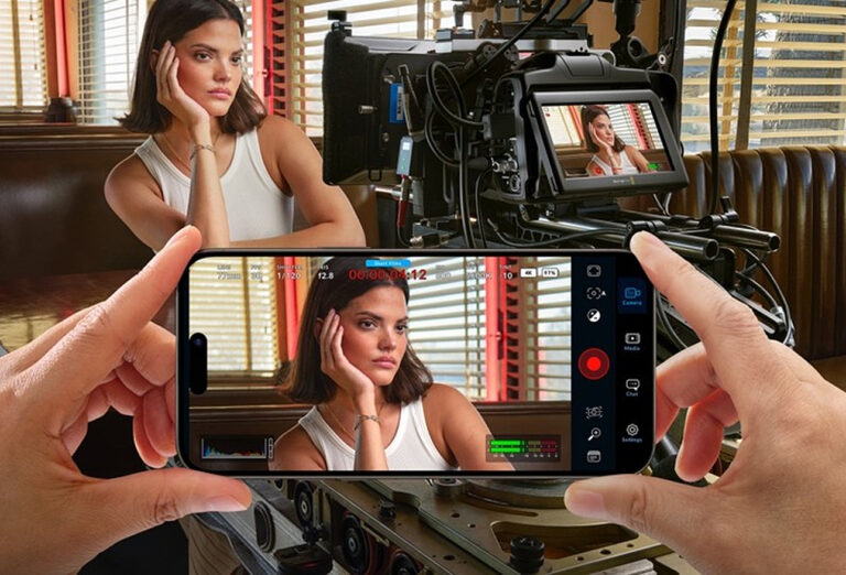 Osoba pozująca przed kamerą, której obraz można zobaczyć na ekranie smartfona trzymanego przez rękę.