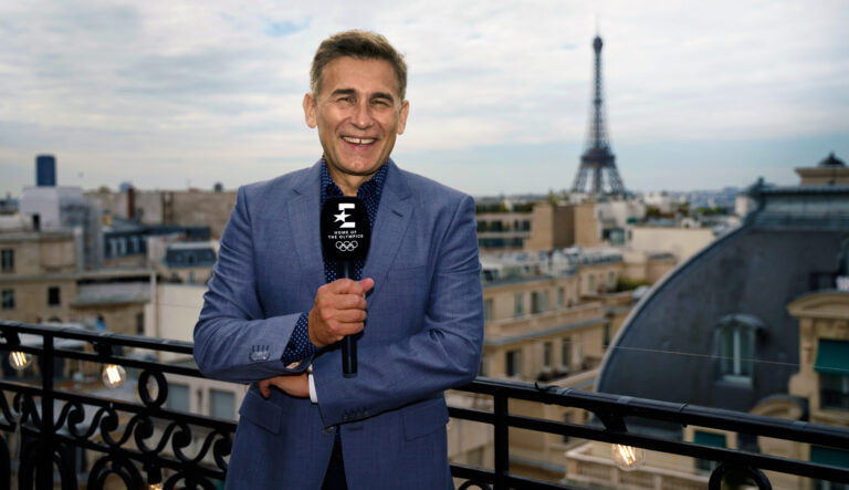 Robert Korzeniowski. Mężczyzna w niebieskiej marynarce trzyma mikrofon na balkonie z widokiem na Wieżę Eiffla w Paryżu.