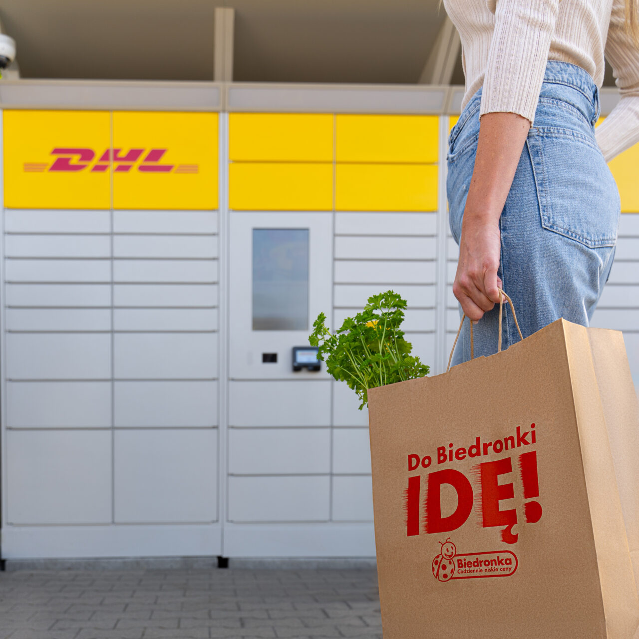 Kobieta trzymająca torbę zakupową z Biedronki przed paczkomatem DHL.