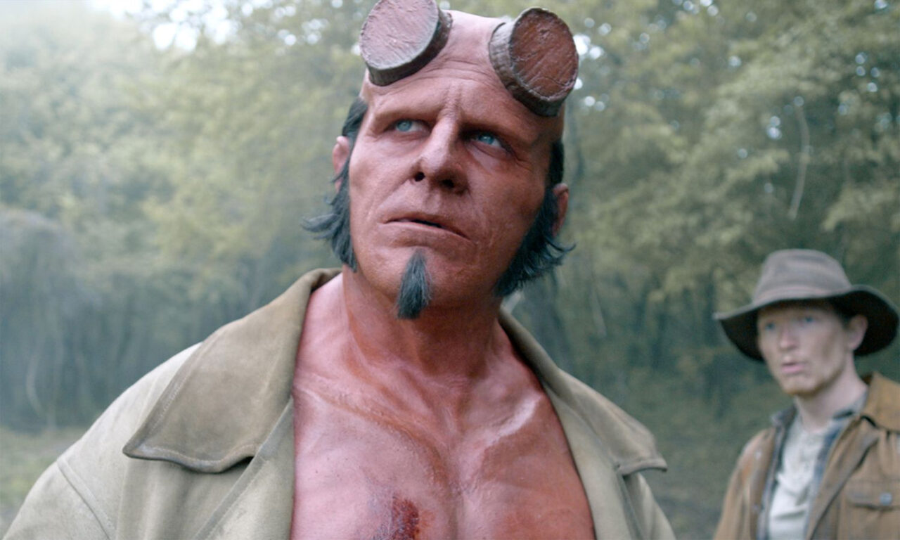 Postać Hellboya z rogami obciętymi do połowy, z brodą, w skórzanym płaszczu, w lesie.